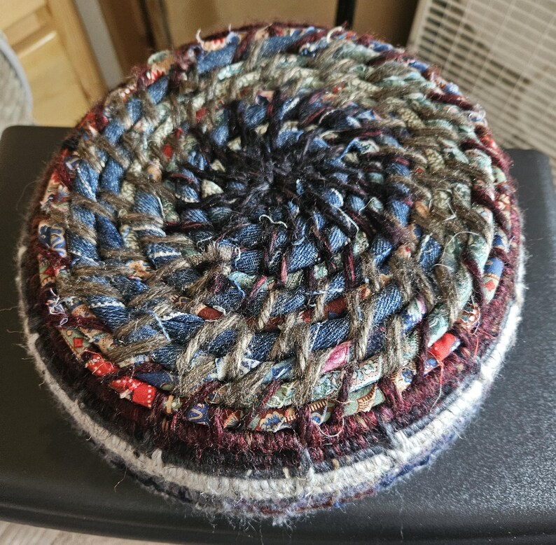 K&ouml;nnte beinhalten: Ein runder, handgefertigter Untersetzer aus geflochtenen Stoffresten. Der Untersetzer hat ein Spiraldesign mit Denim-, roten und gemusterten Stoffstreifen. Die Kanten sind mit einer dunkelburgunderfarbenen und wei&szlig;en Bord&uuml;re versehen. Durchmesser ca. 10 cm.