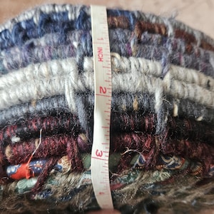 K&ouml;nnte beinhalten: Ein mehrfarbiger, gewebter Textilartikel, etwa 7,6 cm hoch, wird mit einem Ma&szlig;band gemessen. Der Artikel weist eine Mischung aus blauen, grauen, burgunderroten und gr&uuml;nen Farbt&ouml;nen auf. Die Textur erscheint dick und geschichtet, mit sichtbaren Fasern.