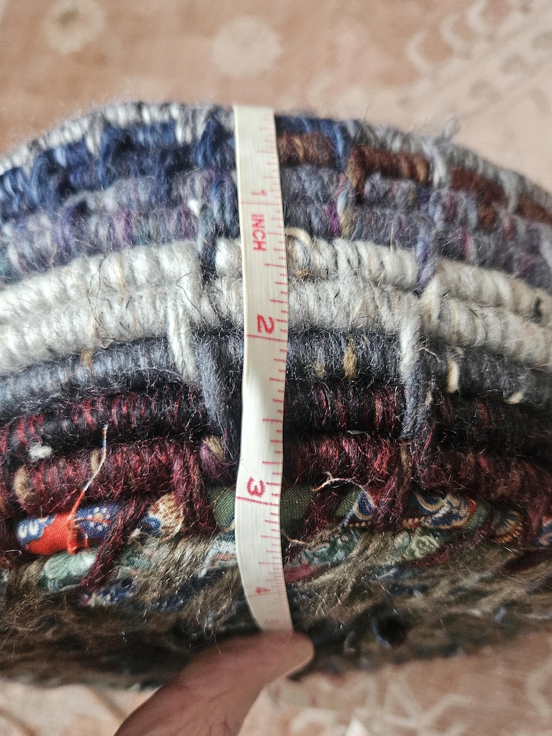 K&ouml;nnte beinhalten: Ein mehrfarbiger, gewebter Textilartikel, etwa 7,6 cm hoch, wird mit einem Ma&szlig;band gemessen. Der Artikel weist eine Mischung aus blauen, grauen, burgunderroten und gr&uuml;nen Farbt&ouml;nen auf. Die Textur erscheint dick und geschichtet, mit sichtbaren Fasern.