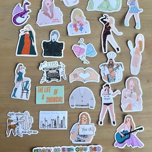 Op de afbeelding: Een verzameling kleurrijke stickers met illustraties van een zangeres, albumhoezen en songreferenties. De stickers bevatten tekst zoals "TS", "Paper Rings", "The Life of a Showgirl", "You ok?" en "You're on your own kid".