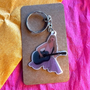 Op de afbeelding: Een sleutelhanger met een zilveren sleutelring, met een persoon die een blauwe gitaar bespeelt. De illustratie staat op een witte achtergrond en draagt een roze jurk. De sleutelhanger is op een bruine kaart gemonteerd.