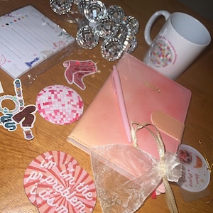 Op de afbeelding: Een verzameling roze en witte briefpapierartikelen, waaronder een notitieboek, een pen en stickers, samen met een witte mok met een kleurrijk ontwerp. Decoratieve discoballichtjes en een cadeauzakje zijn ook aanwezig.