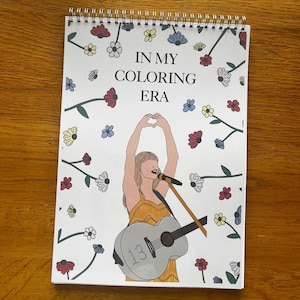 Op de afbeelding: Een spiraalgebonden kleurboek met de tekst "IN MY COLORING ERA" en een tekening van een persoon met een microfoon en gitaar. De omslag is versierd met kleurrijke bloemen en het nummer "13" op de gitaar.