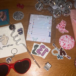 Op de afbeelding: Een verzameling accessoires, waaronder hartvormige zonnebrillen, een notitieblok, stickers en een make-up tasje met de tekst "right where you left me". Andere items zijn een sleutelhanger, discobollichtjes en een roze pen.