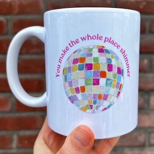Op de afbeelding: Witte keramische mok met een grote handgreep. De mok heeft een kleurrijk discobalontwerp met de tekst "You make the whole place shimmer" in roze. De mok wordt in een hand gehouden.
