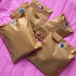 Op de afbeelding: Verschillende bruine papieren enveloppen liggen op een roze ondergrond. Elke envelop is verzegeld met een ronde sticker met een kleurrijk, glinsterend ontwerp. De enveloppen zijn gevouwen en verzegeld, wat suggereert dat ze items bevatten.
