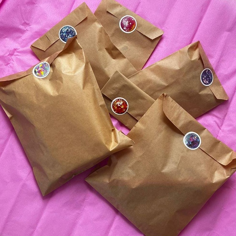 Op de afbeelding: Verschillende bruine papieren enveloppen liggen op een roze ondergrond. Elke envelop is verzegeld met een ronde sticker met een kleurrijk, glinsterend ontwerp. De enveloppen zijn gevouwen en verzegeld, wat suggereert dat ze items bevatten.