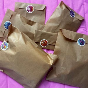 Op de afbeelding: Verschillende bruine papieren pakketten zijn gerangschikt op een roze oppervlak. Elk pakket is verzegeld met een ronde sticker met kleurrijke ontwerpen. De pakketten vari&euml;ren in grootte en zijn licht gekreukt.