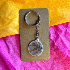 K&ouml;nnte beinhalten: Ein runder Schl&uuml;sselanh&auml;nger mit einem farbenfrohen Mosaikdesign. Der Schl&uuml;sselanh&auml;nger hat einen silbernen Metallring und eine Kette, die an einem transparenten, kreisf&ouml;rmigen Anh&auml;nger mit einem Mosaikmuster aus kleinen, quadratischen Fliesen in verschiedenen Farben befestigt ist.