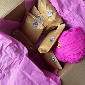 Op de afbeelding: Een kartonnen doos gevuld met bruine papieren pakketten en felroze vloeipapier. De pakketten zijn verzegeld met kleurrijke ronde stickers. De doos is open en toont de inhoud.