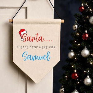 May include: A beige fabric wall hanging banner with a wooden dowel and black string. The text reads Santa in red with a Santa hat icon, followed by PLEASE STOP HERE FOR in black, and the name Samuel in blue script font at the bottom.