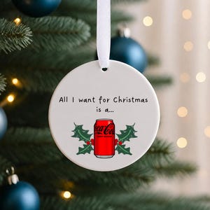 以下が含まれることがあります： 白いリボンで吊るされた丸い白いセラミック製のオーナメント。All I want for Christmas is a...という文字の下に、赤いコカ・コーラ ゼロシュガーの缶のイラストが描かれており、その両側には赤い実をつけた緑のヒイラギの葉が添えられています。