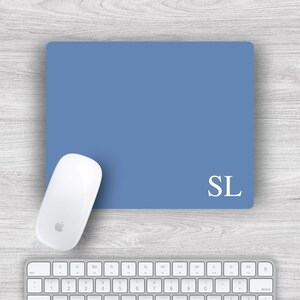 May include: A blue rectangular mousepad with the initials "SL" in white, a white computer mouse, and a white keyboard. The mousepad is on a light wood-grain surface.