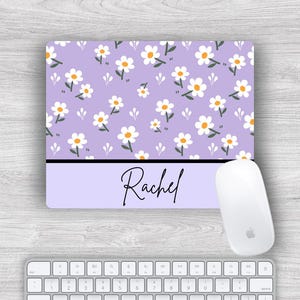 May include: A rectangular mouse mat with a lavender background and a daisy pattern. The mouse mat has a black stripe at the bottom with the name "Rachel" in cursive. A white mouse and keyboard are also visible.