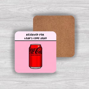 May include: Square coaster with a pink background featuring a red Coca-Cola Zero can. The coaster has the text "RESERVED FOR LEAH'S COKE ZERO" in black. A second, plain brown coaster is visible in the background.
