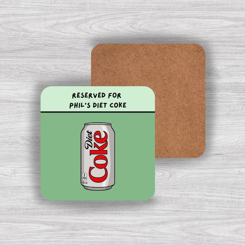 May include: A square coaster with a light green top section featuring the black text RESERVED FOR PHIL'S DIET COKE and a lower section with a graphic of a silver Diet Coke can with red lettering. The coaster has a brown cork backing.