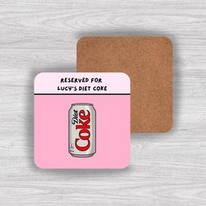 Puede incluir: Un posavasos cuadrado con fondo rosa e ilustración de una lata de Coca-Cola Light. El texto "RESERVED FOR LUCY'S DIET COKE" está impreso en la parte superior. Un segundo posavasos marrón es visible en el fondo.