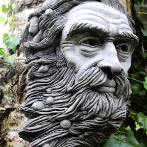 Puede incluir: Una escultura de piedra gris de la cara de un hombre con una larga barba. La barba está esculpida para parecerse a olas y tiene pequeñas conchas incrustadas.