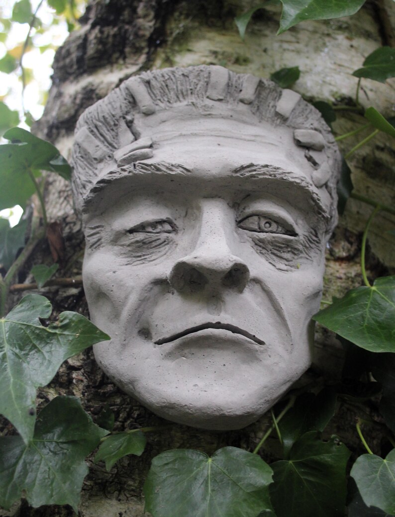 May include: A gray concrete sculpture of a face with a furrowed brow and a serious expression. The sculpture is attached to a tree trunk covered in green ivy.