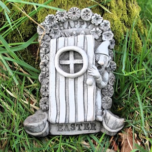 May include: A grey stone Easter garden door with a gnome peeking out. The door features a cross-shaped window and is framed by floral details. The word "HAPPY" is above the door, and "EASTER" is at the base.