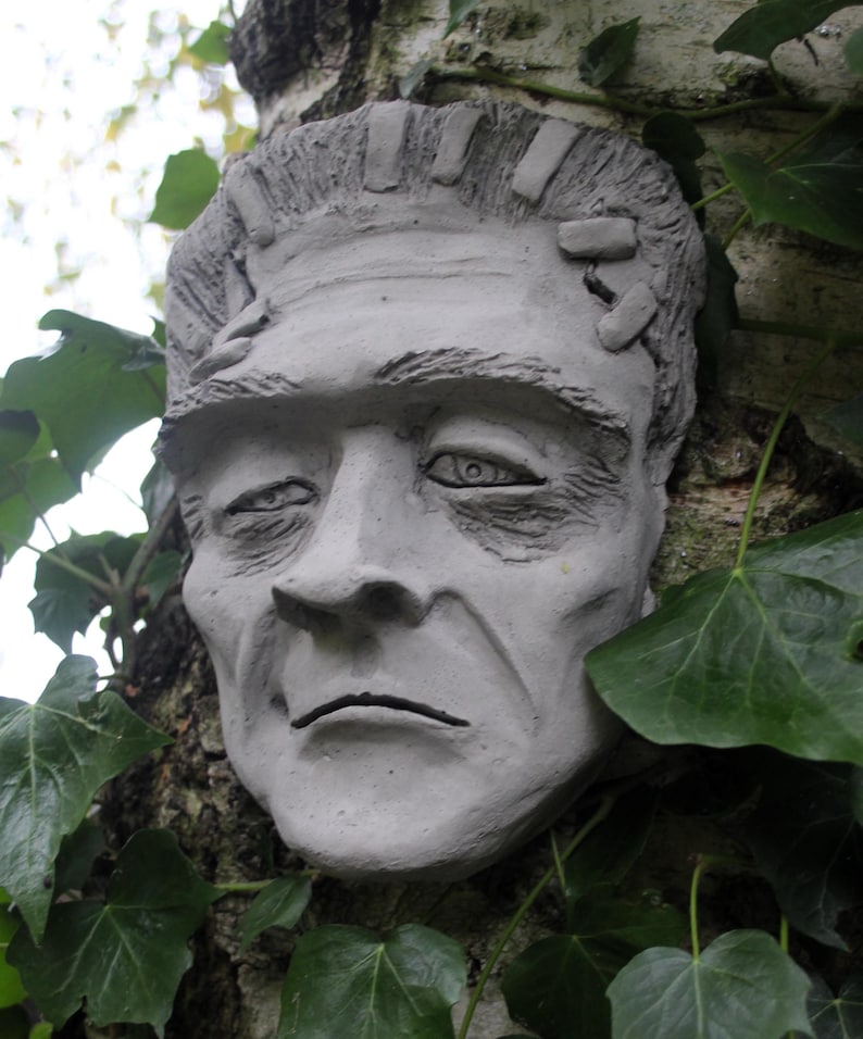 May include: A gray, sculpted face of Frankenstein's monster, with visible stitches on the forehead, is attached to a tree trunk. The face is partially obscured by green ivy leaves.
