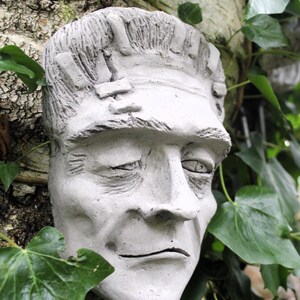 May include: A gray concrete sculpture of a man's face with stitched scars on the forehead. The sculpture is mounted on a tree trunk and surrounded by green ivy.