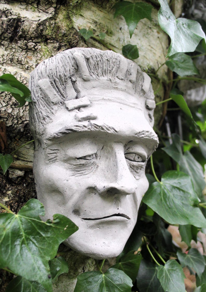 May include: A gray concrete sculpture of a man's face with stitched scars on the forehead. The sculpture is mounted on a tree trunk and surrounded by green ivy.
