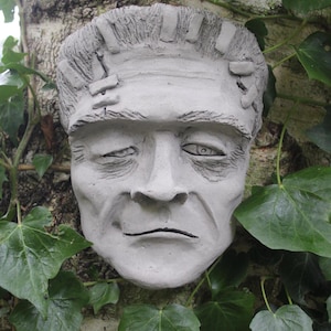 May include: A gray concrete sculpture of a Frankenstein's monster face, with visible stitches on the forehead, set against a backdrop of green ivy.