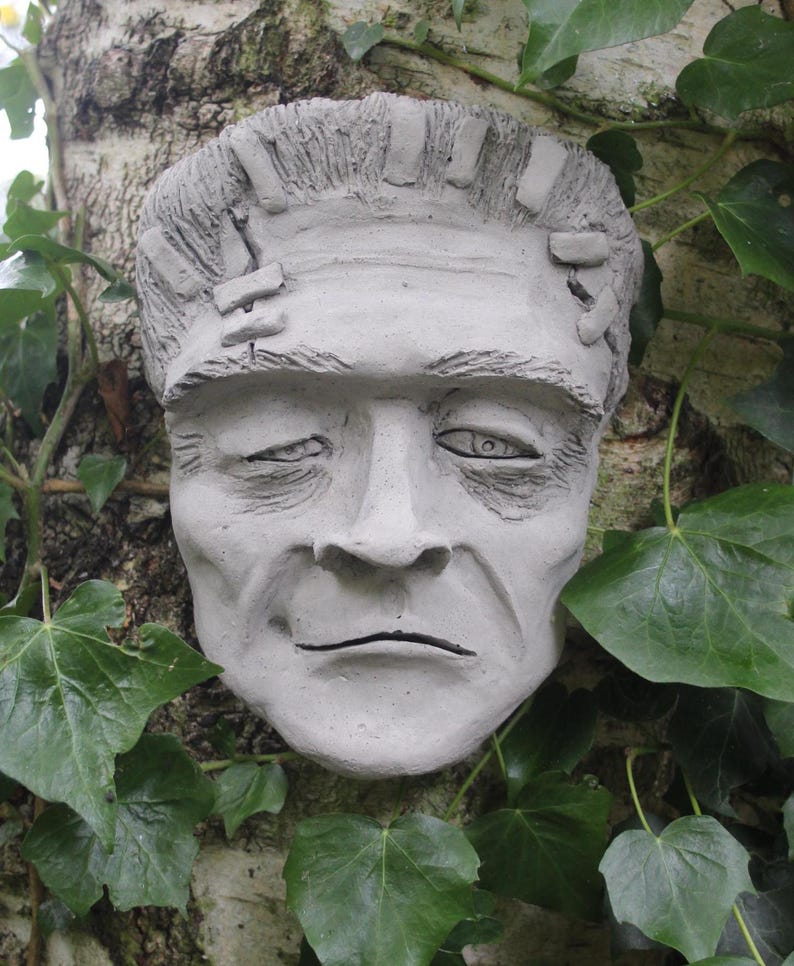 May include: A gray concrete sculpture of a Frankenstein's monster face, with visible stitches on the forehead, set against a backdrop of green ivy.