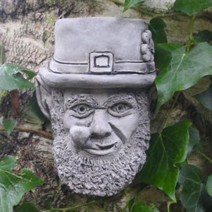 Könnte beinhalten: Eine graue Steinskulptur eines Kobolds mit einem grünen Hut und einem Kleeblatt. Die Skulptur ist an einem mit Efeu bewachsenen Baumstamm befestigt.