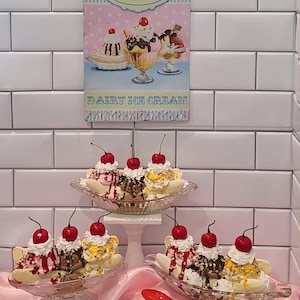 May include: An image of several banana splits in glass dishes, topped with whipped cream and a cherry. The splits are on a pink surface, with a red spoon nearby. A sign that says "The Ice Cream Parlour" is on the wall.