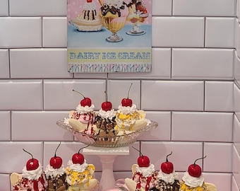 Può includere: Tre coppe di vetro riempite di banana split guarnite con panna montata, ciliegie rosse e vari sciroppi. Un cartello in metallo sopra riporta The Ice Cream Parlour Est. 1901 Dairy Ice Cream. Un cucchiaio di plastica rosso poggia su un tessuto rosa.