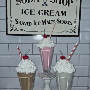 Puede incluir: Tres réplicas decorativas de batidos en vasos de cristal, cada uno con nata montada blanca, una cereza roja y una pajita a rayas. El batido rosa central se asienta sobre un pedestal blanco. Un cartel arriba dice SODA SHOP ICE CREAM SHAVED ICE MALTS SHAKES.