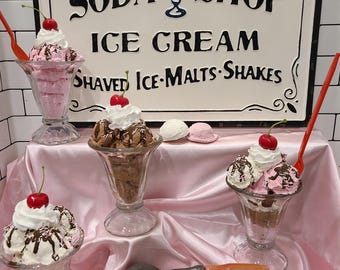 Può includere: Un'insegna vintage di Soda Shop con coppe gelato. L'insegna è bianca con scritte nere e una grafica di gelato blu. Le coppe sono in bicchieri di vetro, guarnite con panna montata, ciliegie e salsa al cioccolato. Una pallina di gelato rosa e una paletta per gelato sono in primo piano.