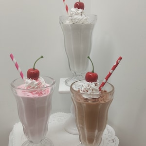 May include: Three milkshakes in classic soda fountain glasses. One is white, one is pink, and one is chocolate. Each is topped with whipped cream, a cherry, and a striped or polka-dot straw. The milkshakes are on a white surface.