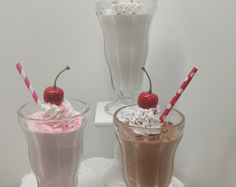 Può includere: Tre frullati in classici bicchieri da fontana di soda. Uno è bianco, uno è rosa e uno è al cioccolato. Ognuno è sormontato da panna montata, una ciliegia e una cannuccia a righe o a pois. I frullati sono su una superficie bianca.