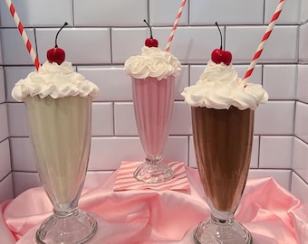 Può includere: Tre frullati in bicchieri alti e trasparenti, ognuno sormontato da panna montata e una ciliegia. Uno è alla vaniglia, uno rosa e uno al cioccolato. Ognuno ha una cannuccia a strisce. I frullati sono su una superficie di raso rosa.
