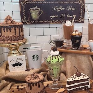 May include: An assortment of desserts and coffee drinks are displayed on a table. A two-tiered chocolate cake sits on a gold stand. Coffee drinks include iced coffee, hot coffee, and a sundae. A sign reads "Coffee Shop open 24 hours."