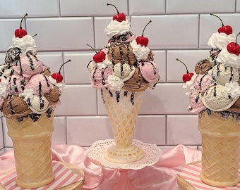 Può includere: Tre sculture decorative a forma di cono gelato con palline rosa, marroni e bianche, guarnite con sciroppo al cioccolato, panna montata bianca e ciliegie rosse. Ogni cono presenta una trama a cialda ed è posizionato in verticale.