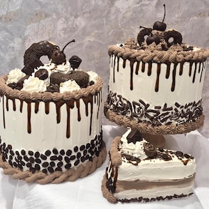 May include: Two round cakes and one triangular cake slice featuring white frosting, dark chocolate ganache drips, chocolate shavings, coffee beans, whipped cream dollops, chocolate donuts, and cherries on top of the whole cakes.