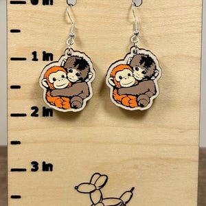 May include: A pair of dangle earrings featuring a cartoon illustration of two hugging monkeys. The earrings are orange, brown, and black, with silver-colored hooks. The earrings are displayed on a wooden stand with a balloon dog illustration.