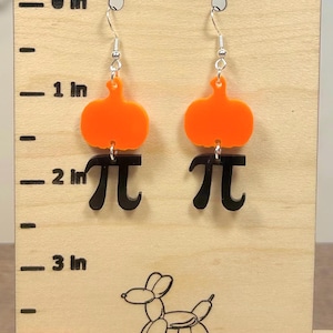 May include: A pair of orange pumpkin-shaped earrings with black pi symbols below. The earrings hang from silver hooks. The earrings are displayed on a wooden stand with inch measurements.