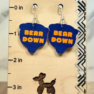 May include: A pair of blue, bear-shaped earrings with the orange text 'BEAR DOWN' printed on each. The earrings are displayed on a light-colored wooden stand with a ruler marking up to 3 inches and the text 'BALLOON DOG BOUTIQUE' at the bottom.