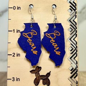 May include: A pair of blue, state-shaped dangle earrings featuring the orange text 'Bears' displayed on a wooden display card with a ruler marked in inches, showing the earrings measure approximately 2 inches in length from the top of the hook.