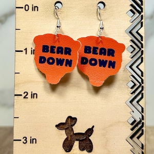 May include: A pair of orange leather earrings with the text BEAR DOWN in navy blue, hanging from silver-toned hooks on a wooden display card with a ruler showing a length of approximately 1.5 inches for the earrings.