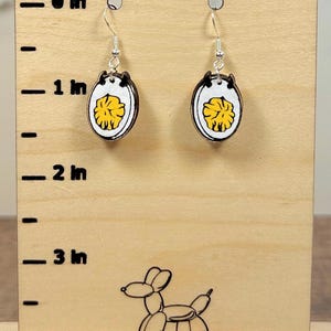 May include: A pair of dangle earrings featuring a fried egg design with a white oval border and a yellow yolk center. The earrings are displayed on a wooden stand with a ruler marking 0 to 3 inches, showing the charms are approximately 0.5 inches.