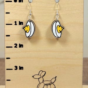 May include: A pair of dangle earrings featuring a cartoon-style fried egg design. The earrings have a white background with a yellow yolk and are displayed on a wooden stand with inch measurements.