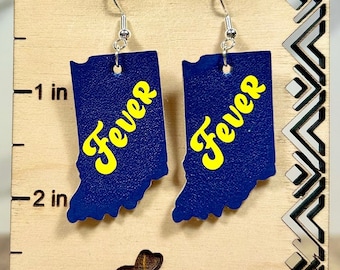 May include: A pair of blue dangle earrings shaped like the state of Indiana, featuring the word Fever in yellow script. The earrings are displayed on a light wood stand with a ruler marking up to 3 inches and the text Balloon Dog Boutique.