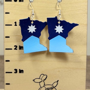 May include: A pair of earrings shaped like the state of Minnesota. The top portion is navy blue, the bottom is light blue, and a white star is in the center. The earrings hang from silver hooks.