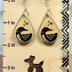 May include: A pair of teardrop-shaped wooden earrings featuring a laser-engraved loon and star design. The earrings hang from silver-toned metal hooks on a light-colored wooden display card marked with a 3-inch ruler and the text Balloon Dog Boutique.